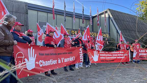 Des membres d'Unia se tiennent devant la Halle des Expositions à Delémont avec des drapeaux et des banderoles exigeant de meilleures conditions de travail dans le secteur de la construction.